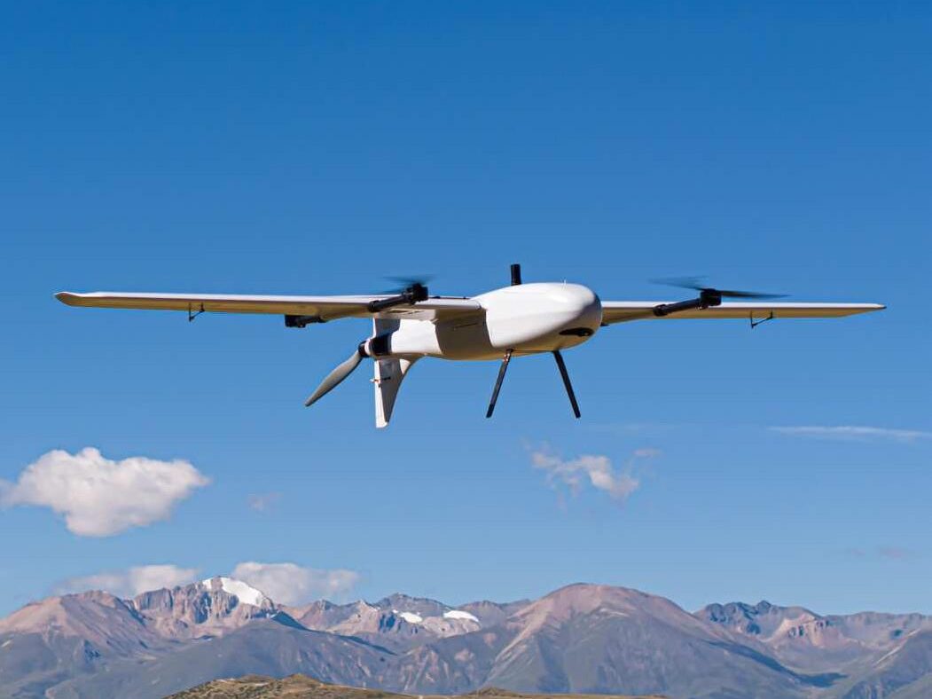 White SA240 Pro fixed-wing VTOL drone flying under a clear blue sky, with its four lift rotors and rear pusher propeller visible